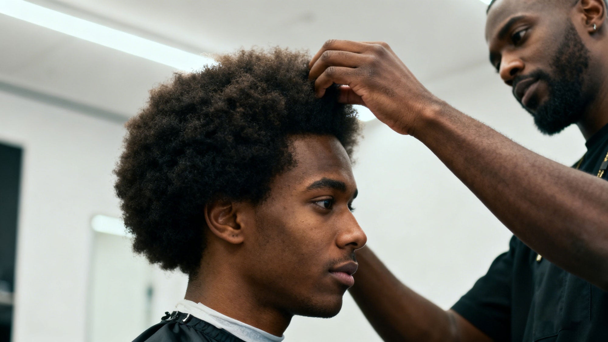 A young Black man with an afro hairstyle getting his hair styled by a barber in a salon.